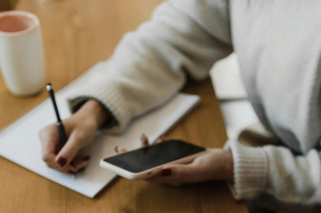 Una persona con un suéter blanco consulta un teléfono móvil mientras escribe en un notepad.